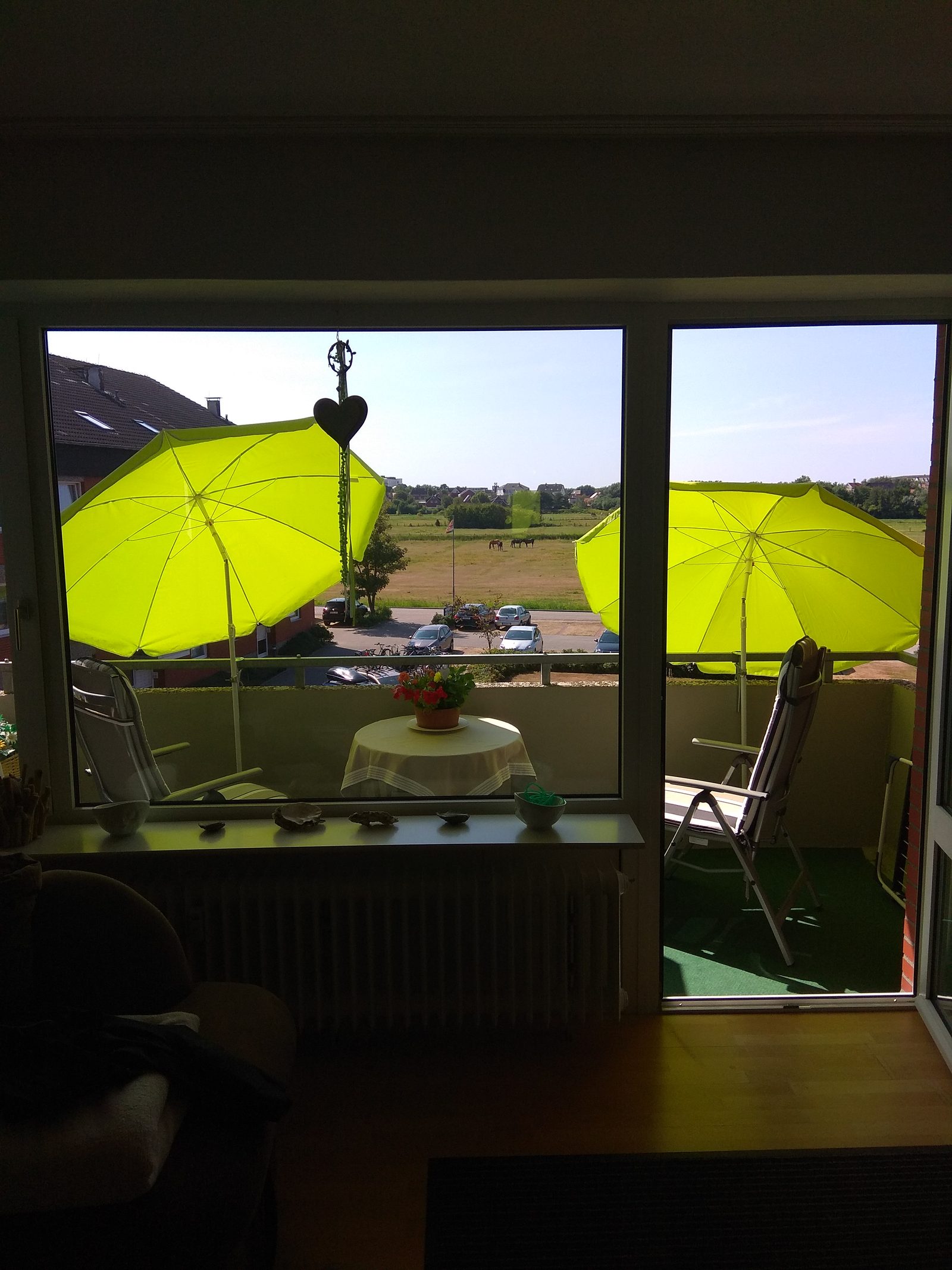 Blick aus dem Wohnraum durch das Fenster auf den Balkon mit Sonnenschirmen und Pferdewiese